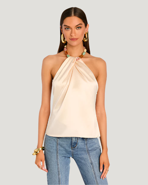 A model wearing a light beige strapless halter top with a pleated design at the neckline. The halter top includes a colorful beaded necklace as part of its design. The beads are turquoise, pink, and beige shades. The top is paired with high-waisted blue jeans.  The model is also wearing a chunky bracelet and is placed behind a blank background.