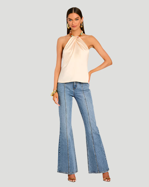 A model wearing a light beige strapless halter top with a pleated design at the neckline. The halter top includes a colorful beaded necklace as part of its design. The beads are turquoise, pink, and beige shades. The top is paired with high-waisted blue jeans and high heels. The model is also wearing a chunky bracelet and is placed behind a blank background.