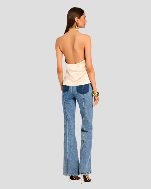The backside view of a model wearing a light beige strapless halter top with a pleated design at the neckline. The top is paired with high-waisted blue jeans and high heels. The model is also wearing a chunky bracelet and is placed behind a blank background.