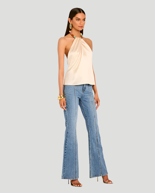 A model wearing a light beige strapless halter top with a pleated design at the neckline. The halter top includes a colorful beaded necklace as part of its design. The beads are turquoise, pink, and beige shades. The top is paired with high-waisted blue jeans and high heels. The model is also wearing a chunky bracelet and is placed behind a blank background.