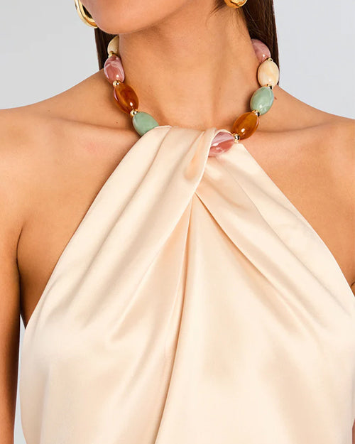 A close-up frontal view of a model wearing a light beige strapless halter top with a pleated design at the neckline. The halter top includes a colorful beaded necklace as part of its design. The beads are turquoise, pink, and beige shades.