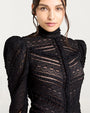 Close up of a person wearing a black lace shirt with puffed sleeves and semi-sheer striped detailing, against a white background