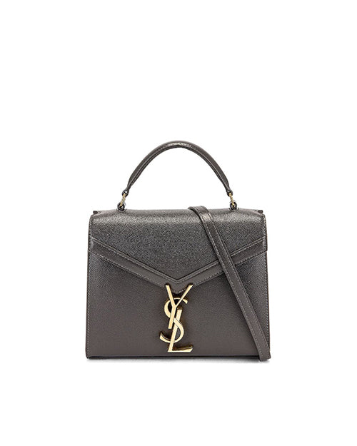 A structured, charcoal grey designer handbag with a prominent gold-toned metal logo in the shape of the letters ‘YSL’ intertwined on the flap. The bag features a top handle and a visible shoulder strap, suggesting versatility in carrying options.