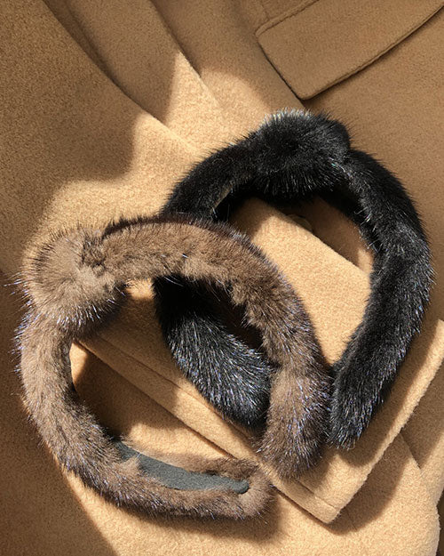 Two fur headbands are placed on a beige fabric surface. The headband on the left is black, and the one on the right is brown with natural tones.