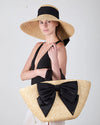 Woman wearing a straw hat with a black ribbon and holding a matching straw bag with a large black bow on a white background.