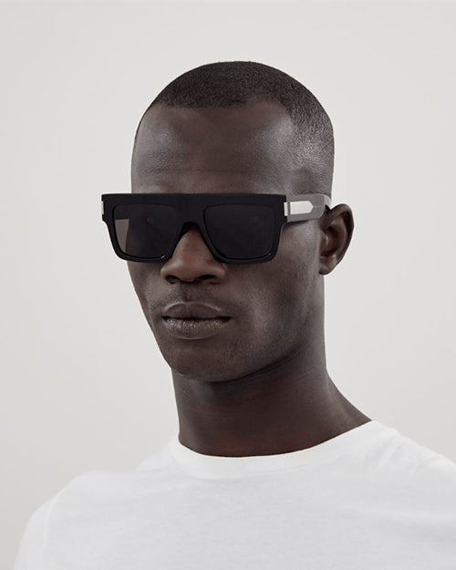 Person wearing a pair of black Saint Laurent sunglasses with a thick frame and large, dark lenses.  The design is sleek and modern, suggesting a fashionable accessory.
