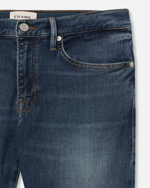 A close-up image of the upper part of a pair of dark blue denim jeans. The jeans feature a visible brand label marked ‘FRAME’ on the waistband, a metal silver button closure, and rivets at the pocket corners. The stitching is in a contrasting orange color, and there is a small watch pocket within the right front pocket. The fabric has subtle variations in color, typical of denim material.