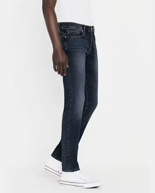 A side view of a pair of dark blue denim jeans with a faded effect around the thigh and knee area. The jeans have a fitted cut, visible stitching, and are styled with a black belt, white shirt and white sneakers.