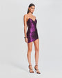 A side view of a strapless, shiny purple mini dress with ruched detailing and a sweetheart neckline. The individual is also holding a black clutch and wearing open-toed black heels. 