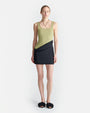 A full body view of a blonde model wearing a dress with an asymmetrical black mini skirt design and a green tank top with a square neckline. The model is wearing chunky black slides, a black necklace, and is standing against a white background.