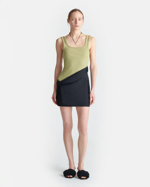 A full body view of a blonde model wearing a dress with an asymmetrical black mini skirt design and a green tank top with a square neckline. The model is wearing chunky black slides, a black necklace, and is standing against a white background.