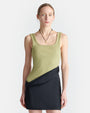 A close-up view of a blonde model wearing a dress with an asymmetrical black mini skirt design and a green tank top with a square neckline. The model is also wearing black necklace with a small gold pendant, and is standing against a white background.