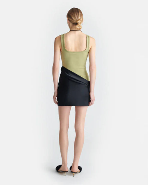 The backside view of a blonde model in a bun wearing a dress with an asymmetrical black mini skirt and a green tank top with a low, square neckline. The model is wearing chunky black slides, a black necklace, and is standing against a white background.