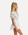 A sideview of a blonde model wearing a white dress with short sleeves and ruffled details on the shoulders, down the sides of the waist, and the skirt with a layered design. The background is plain white.