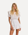 A knee-up view of a blonde model wearing a white dress with short sleeves and ruffled details on the shoulders, down the sides of the waist, and the skirt with a layered design. The chest area has vertical lace detailing. The background is plain white.