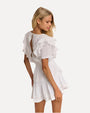 A knee-up backside view of a blonde model wearing a white dress with short sleeves and ruffled details on the shoulders, down the sides of the waist, and the skirt with a layered design. The back has a slit down the middle. The background is plain white.