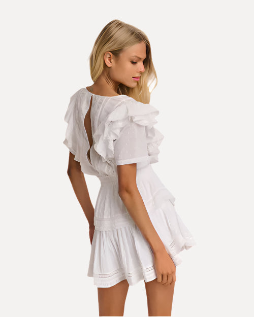 A knee-up backside view of a blonde model wearing a white dress with short sleeves and ruffled details on the shoulders, down the sides of the waist, and the skirt with a layered design. The back has a slit down the middle. The background is plain white.