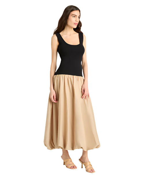 Front view of a  person standing in front of a plain background wearing a sleeveless dress with a black bodice and a voluminous, ankle-length beige skirt. They are also wearing beige open-toe sandals.