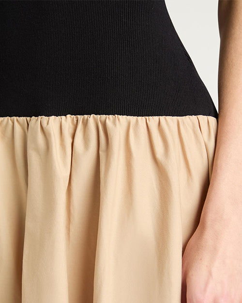 Close-up of a person wearing a dress with a fitted black top and a gathered beige skirt. The person's right hand rests against the beige fabric.