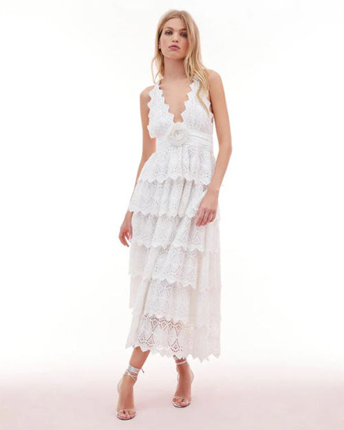 Model wearing a detailed white halter dress with multiple layers and patterns, paired with light-colored sandals, posed against a plain background.