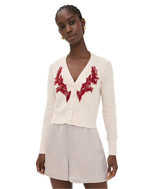 Woman wearing a cream cardigan with red floral embroidery and nude shorts on a white background
