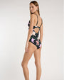 Back view of a person standing against a light neutral background, showing a black floral one piece swimsuit with thin spaghetti straps, a V-neckline, and an underbust seam. The fabric features pink and lavender floral prints with green stems and leaves.
