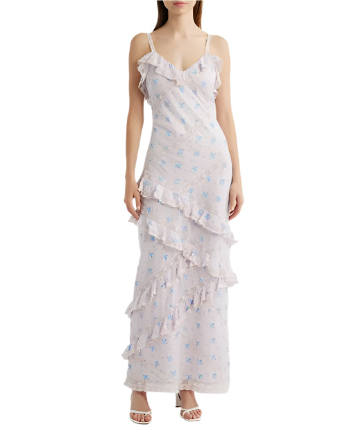 A person is seen wearing a long, sleeveless dress with thin straps. The dress is white with a subtle blue floral pattern and features ruffles along the neckline, across the front leg area, and at the hem. The person is also wearing white open-toed shoes.