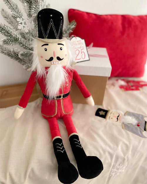 A festive nutcracker doll in a red outfit sits on a beige surface next to a gift box marked "25" and a red pillow, conveying a Christmas theme.