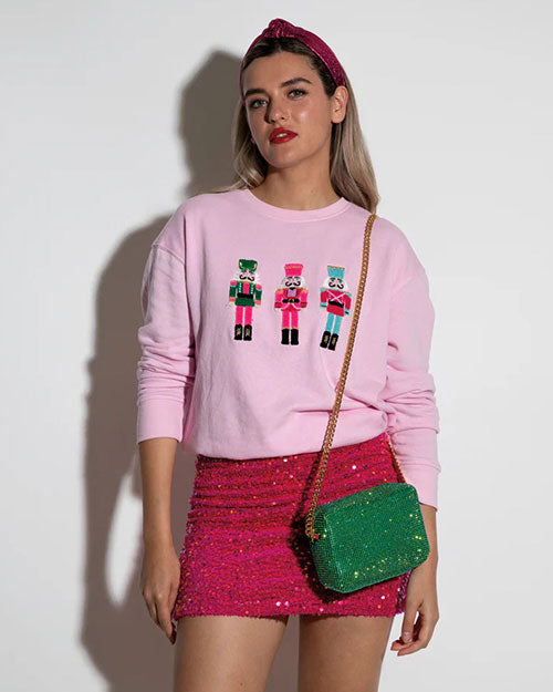 A person stands against a light-colored background, wearing a pink long-sleeved sweatshirt with graphic designs of three nutcracker on the front, paired with a sparkling pink mini skirt. They have a green crossbody bag with studs and a headband.