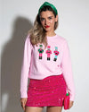 A person stands against a light-colored background, wearing a pink long-sleeved sweatshirt with graphic designs of three nutcracker on the front, paired with a sparkling pink mini skirt.