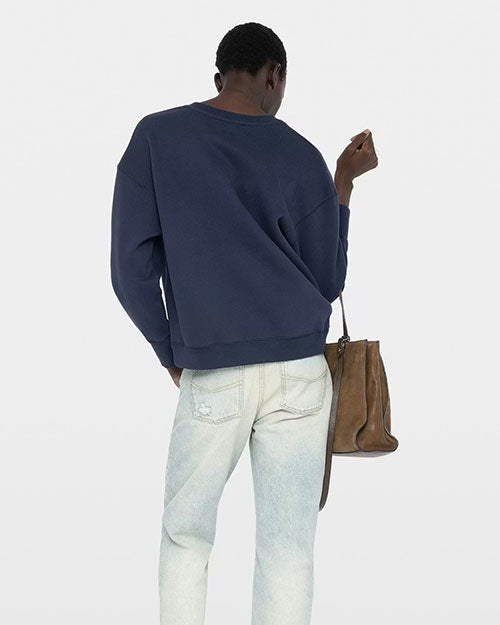 A person is shown from the back wearing a navy blue sweatshirt with a relaxed fit and long sleeves. The sweatshirt has a simple design with no visible graphics on the back. The individual is also wearing light‑wash jeans with slight distressing on one pocket. In one hand, the individual holds a brown leather handbag with a structured shape and a soft, slouchy appearance. The background is plain and light-colored.