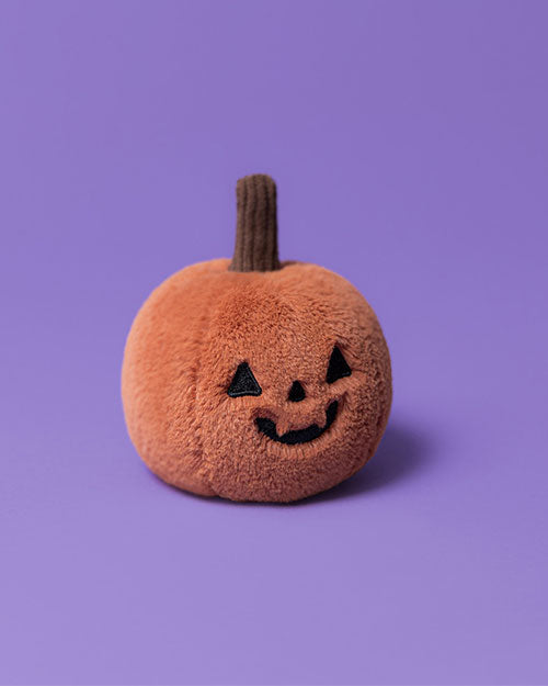 Orange plush pumpkin toy with a brown stem, black triangular eyes, and a smiling jack-o'-lantern face, set against a purple background.