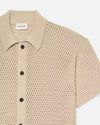 A close-up image of a beige, short-sleeved, knitted polo shirt with a ribbed collar. The garment features a visible texture from the knit pattern and has a row of dark buttons down the front. 