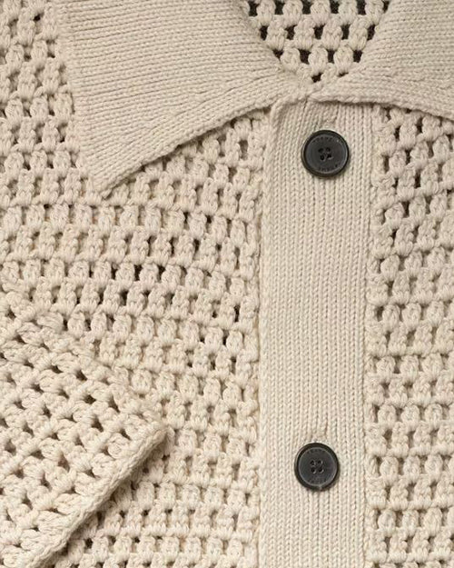 Close-up of a beige, short-sleeved, knitted polo shirt with a ribbed collar. The garment features a knit pattern and has a row of dark buttons down the front.