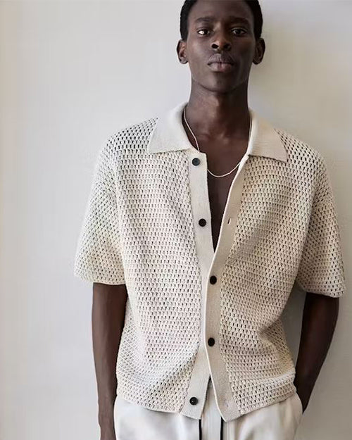 Model wearing a beige, short-sleeved, knitted polo shirt with a ribbed collar. The garment features a knit pattern and has a row of dark buttons down the front. The model styles top with beige shorts.