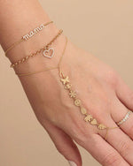 A close-up image of a person’s hand against a beige background, adorned with three delicate gold bracelets. The bracelets feature various charms including the word ‘mama’, a heart outline, and a sequence of butterfly and flower shapes.