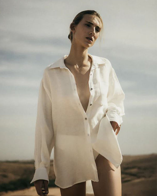 A model stands in a desert landscape, wearing an unbuttoned white shirt.