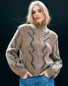 Person wearing a brown knitted sweater with a high collar, featuring vertical wavy lines and leaf-like patterns, paired with blue jeans.