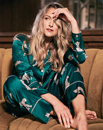 A person is seated on a brown sofa wearing an emerald green silk pajama set  adorned with a white leopard print pattern. The setting appears to be a cozy indoor environment.