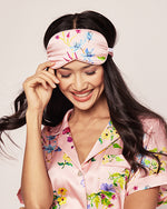 A model wearing a pink sleeping mask featuring blue, yellow, and pink flowers. The mask has a ruched elastic band for securing around the head. The model is wearing a matching pink pajama top.