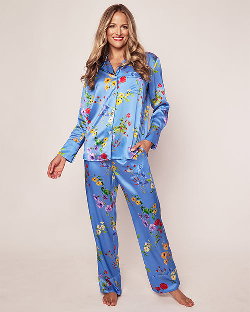 A model wearing a satin blue pajama set with flowers in shades of pink, yellow, green, and red. The top is a button-up with a notched collar, and the matching pants have a relaxed fit. 