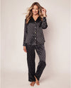 A model in a black silk pajama set with a long-sleeved button-up shirt and matching pants. The set features an all-over print of small white motifs. The shirt has a notched collar, a pocket on the left breast.