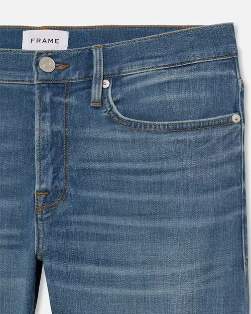 Close-up of a pair of blue denim jeans focusing on the upper left section, showcasing the waistband with a visible brand label marked ‘FRAME,’ a metal button, and rivets. The fabric has varying shades of blue with faded areas and distinct wrinkle details suggesting stretchy material.