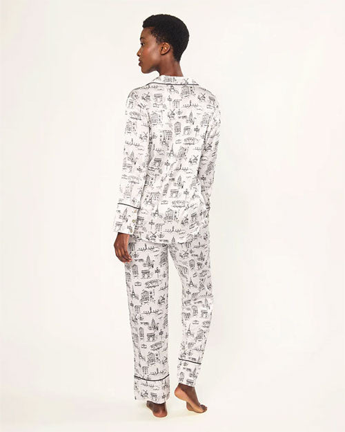  Person standing barefoot with their back to the camera, wearing a white pajama set with black printed illustrations of buildings and trees. The set includes a long-sleeved, button-down top and full-length pants; one leg is slightly raised.