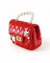 A side view of a small, red quilted handbag with a pearl handle. The bag features a gold clasp in the center and is adorned with white flower appliques, each with a gold center, distributed evenly.