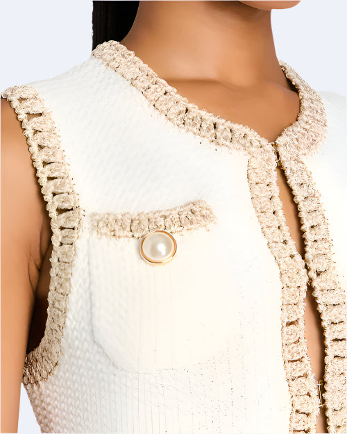 A close-up frontal image of a person wearing a white textured sleeveless top with beige trim. The fabric of the top has a textured appearance, which adds depth and interest to the design. The texture is created by small, raised patterns that give the fabric a slightly bumpy feel. The top has a decorative pocket on the left side, adorned with a large pearl-like button encircled by a gold ring.