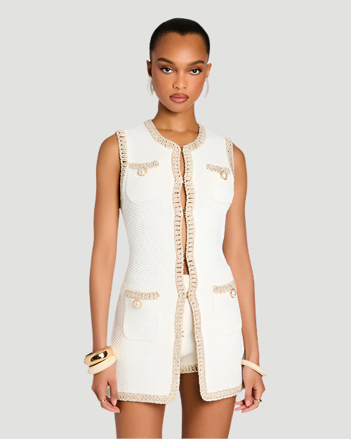 A person wearing a sleeveless white vest with beige trim and buttons. The fabric of the dress appears to be lightweight and breathable. There are two chest pockets and two hip pockets, all with button closures. The chest pockets are positioned just below the shoulders, and the hip pockets are placed near the waist. The beige trim runs along the edges of the dress, including the neckline, armholes, and pocket flaps. The person is accessorizing with two white bracelets on.