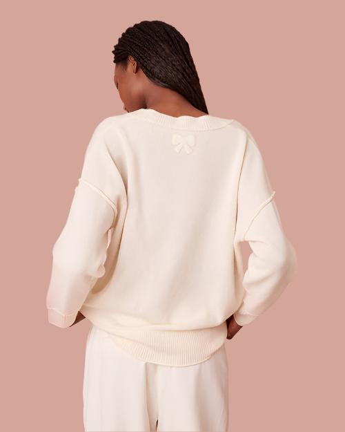 A back view of a cream-colored sweater with a ribbed hem and cuffs. The sweater features a unique detail of a small, textured bow at the nape of the neck. Th sweater features purposeful topstitching.