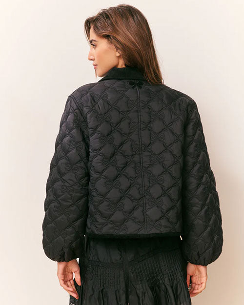 Back view of a black quilted jacket featuring a diamond-pattern stitch design, long sleeves, and a straight hem, paired with a black mini skirt.