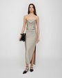 A person standing against a plain background wearing a light taupe, satin‑finish midi dress with thin spaghetti straps. The dress features a draped neckline decorated with a row of hanging bead-like embellishments and a high side slit that extends above the knee. The individual is holding a structured black clutch bag in one hand and wearing pointed, strappy flat shoes. The overall silhouette is fitted and smooth, with the hem falling to mid‑calf.